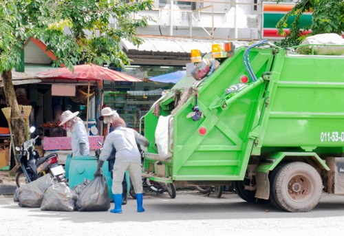 A green waste collection vehicle parked on a street during daylight hours, with two workers in white uniforms and protective gloves handling black garbage bags filled with rubbish. The vehicle has a hydraulic lifting mechanism at the rear, and the scene is set in an urban area with storefronts, trees, and a paved sidewalk visible in the background. The workers are positioned in front of the truck, actively loading or unloading waste, with one worker bending down and the other standing nearby. Surrounding the scene are parked motorcycles and some outdoor market stalls with umbrellas, indicating a busy local environment. The scene is well-lit by natural sunlight, emphasizing the textures of the plastic bags, the metal surface of the recycling truck, and the various elements of the street scene, suggesting the type of rubbish removal service provided by Office Clearance Brixton in the Brixton area, post code SW9.