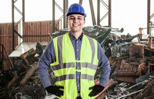 A young male worker in a high-vis yellow safety vest, blue shirt, and blue safety helmet standing inside an industrial or warehouse space filled with a large pile of assorted rubbish and debris. The debris includes broken wooden pallets, metal sheets, and various discarded materials, some of which appear rusted or damaged. The background features metal structural beams supporting the building’s roof and windows allowing natural light to illuminate the scene. The worker is smiling confidently, with gloved hands on his hips, suggesting a professional rubbish removal or clearance operation, possibly managed by Office Clearance Brixton in the Brixton area, close to postcode SW9. The environment indicates a site prepared for rubbish collection or clearance, emphasizing the importance of safety gear and organized waste management in urban or industrial settings.