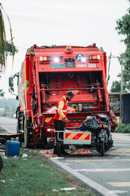 An outdoor scene showing a large pile of mixed household and office waste accumulated around several recycling bins and trash containers on a paved pavement near a commercial building. The waste inclu