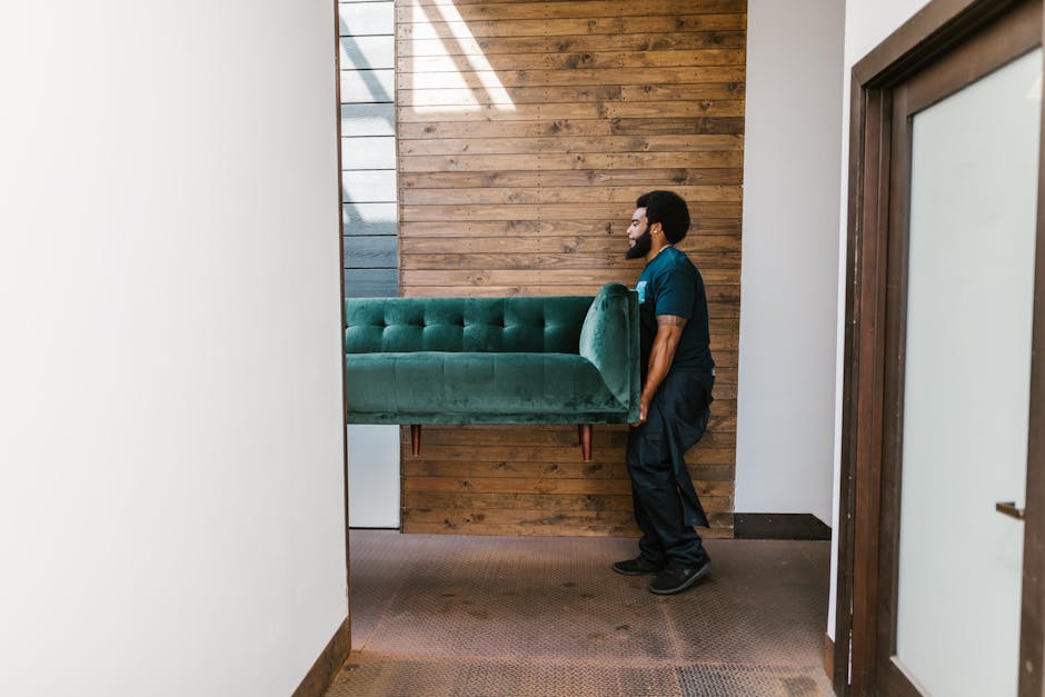 A man with a dark complexion, curly black hair and beard, dressed in a dark blue T-shirt and matching trousers, is carrying a vintage-style armchair with a teal-green upholstered fabric and wooden car